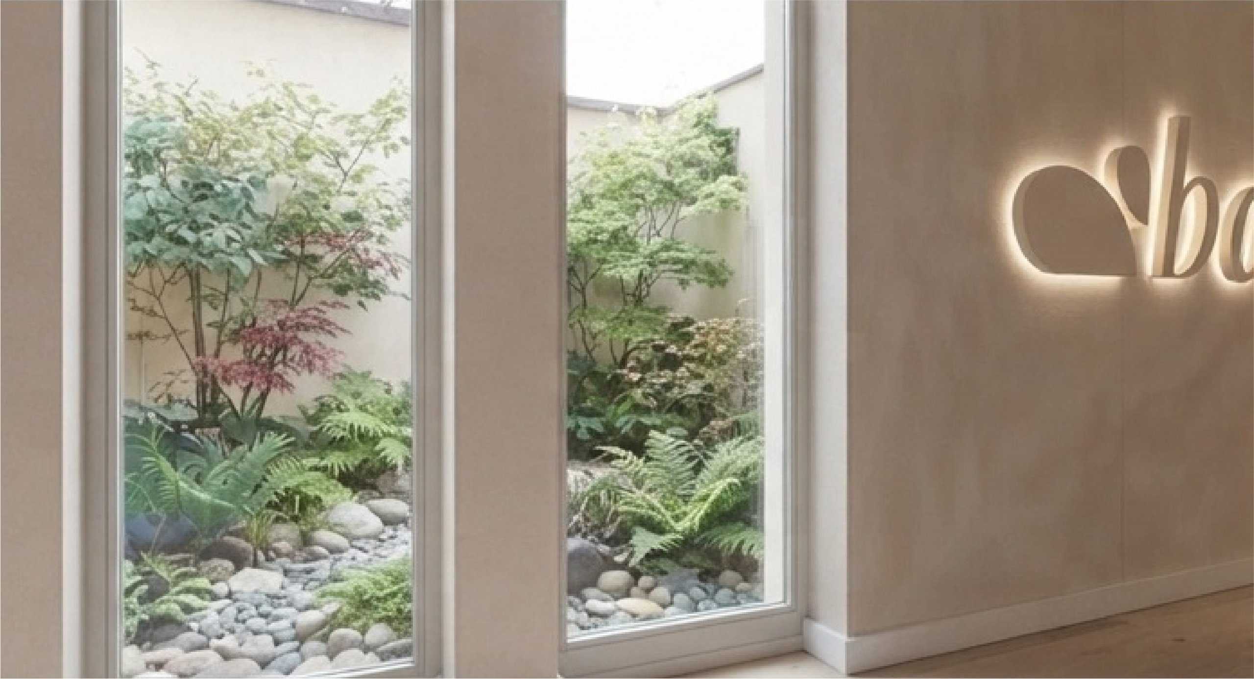 Interior hallway with tall windows overlooking a lush stone-filled courtyard and a glowing partial wall sign.